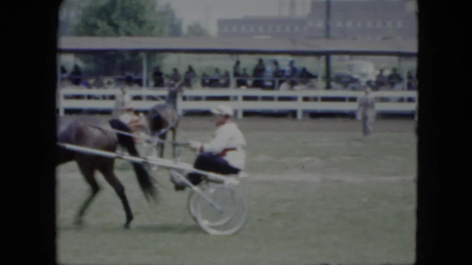 1951 Cleveland fairground frame with a harness horse, sulky driver, white railings, spectators, trees, and a building in the background.