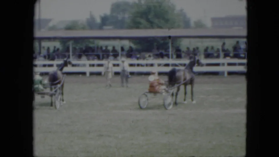 Two sulky teams appear on the grassy Cleveland arena, with one closer to camera and spectators under a covered stand.