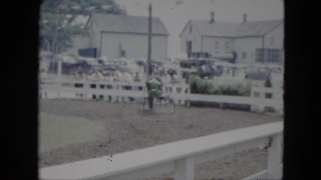 The horse-drawn racing cart continues around the Cleveland dirt track, seen through soft 8mm color with parked cars and buildings in back.