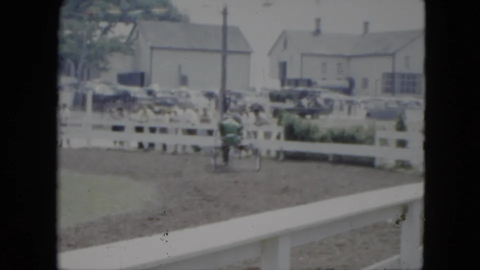 The horse-drawn racing cart continues around the Cleveland dirt track, seen through soft 8mm color with parked cars and buildings in back.