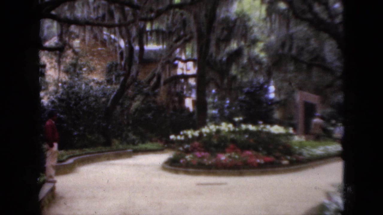 A curved garden path passes flower beds and moss-draped trees with a visitor partly visible.