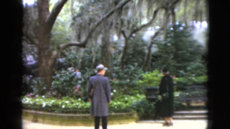 1952 Florida home-movie frame of a man in a coat and a woman with an umbrella walking through a planted garden.
