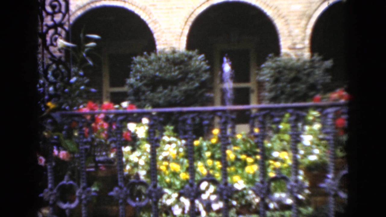 1952 Florida 8mm view of a courtyard garden with arched brick openings, wrought-iron railing, flowering plants, shrubs, and a fountain spray.