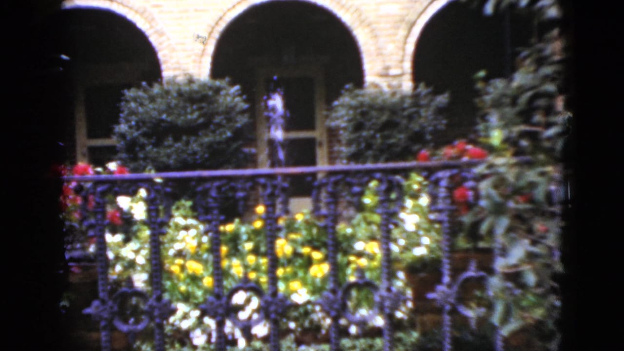 1952 Florida hotel courtyard view with flowers, trimmed shrubs, iron railing, arched brick architecture, and a small fountain at center.