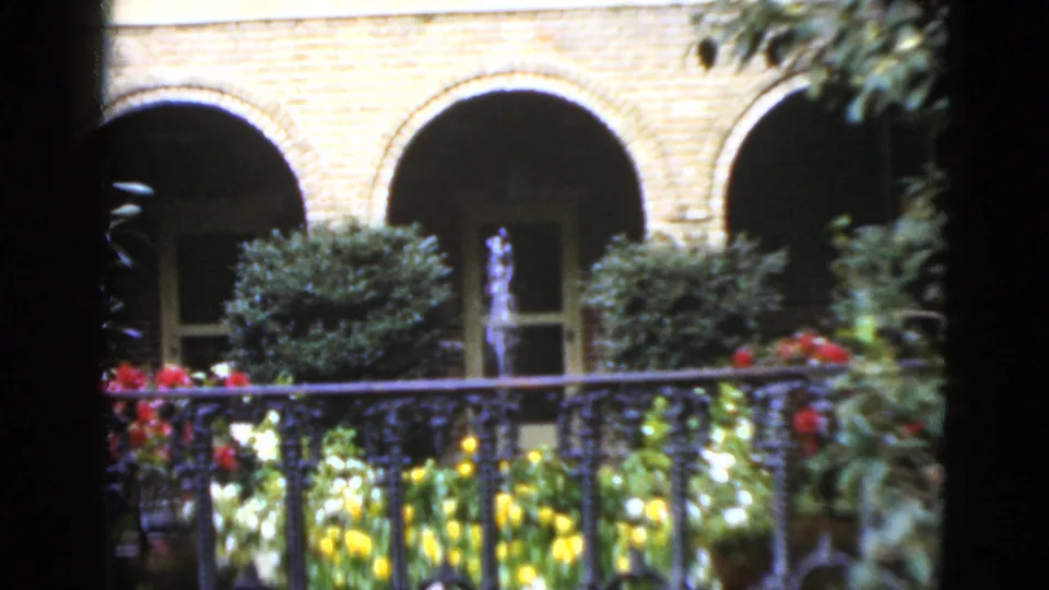 1952 Florida 8mm frame showing a courtyard garden through ironwork, with arched brick openings, bright flowers, shrubs, and a central fountain.
