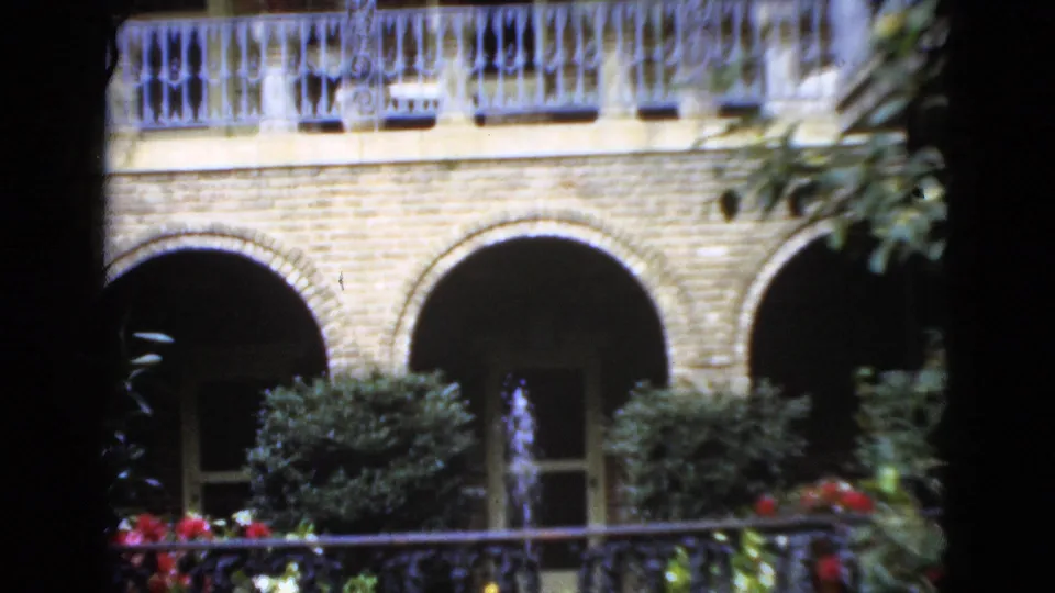 1952 Florida 8mm view of a courtyard facade with upper balcony rail, brick arches, potted-looking shrubs, flowers, and a small fountain.