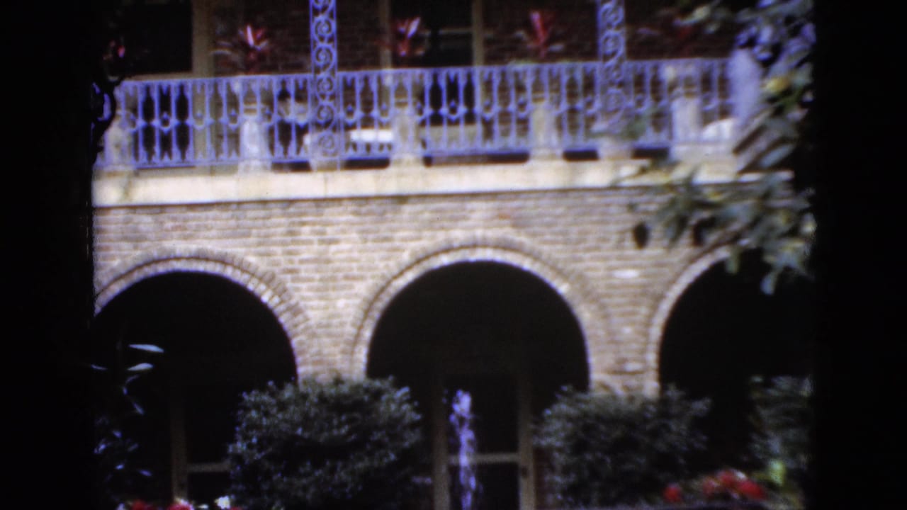 1952 Florida 8mm frame showing a brick building with arched openings, ornate balcony rail, potted plants, and shadowed foliage framing the view.