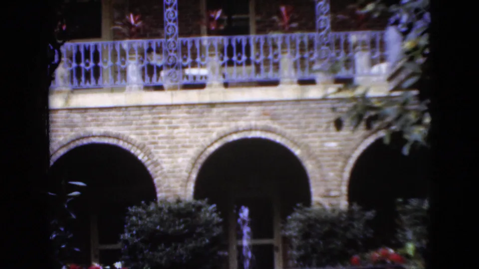 1952 Florida 8mm frame showing a brick building with arched openings, ornate balcony rail, potted plants, and shadowed foliage framing the view.