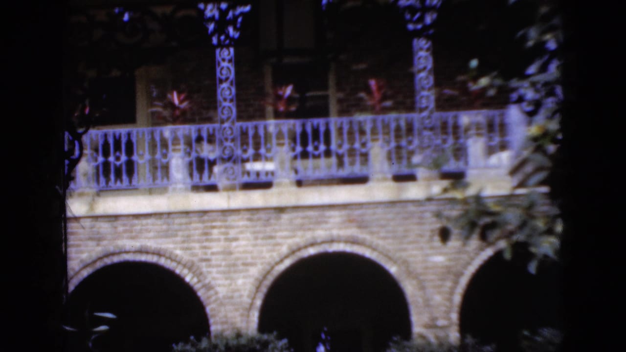 1952 Florida home-movie frame of a brick arcade and decorative balcony with plants, viewed through dark leaves and branches.