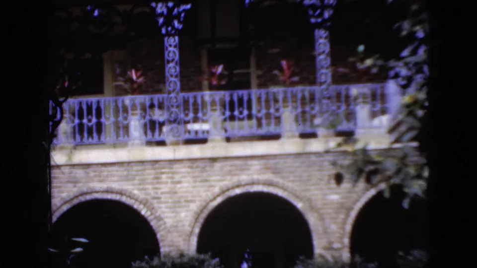 1952 Florida home-movie frame of a brick arcade and decorative balcony with plants, viewed through dark leaves and branches.