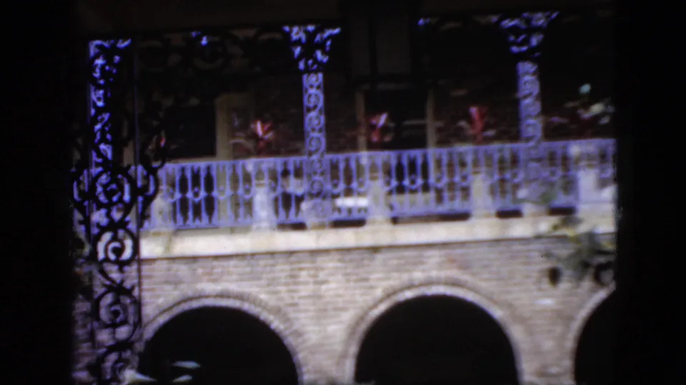 1952 Florida 8mm frame with brick archways, decorative balcony ironwork, potted plants, and dark foliage across the lower foreground.
