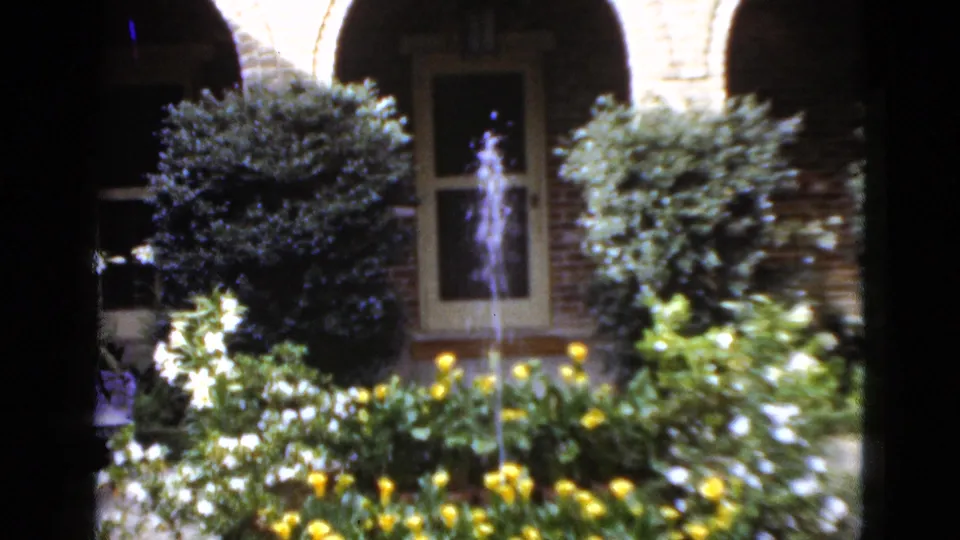 1952 Florida 8mm frame showing a landscaped courtyard with shrubs, flowers, an arched brick entry, doorway, and a small fountain spraying water.
