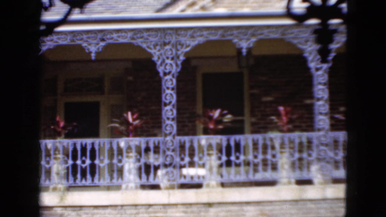 1952 Florida 8mm frame showing white decorative balcony ironwork, brick walls, dark porch openings, and potted red foliage.