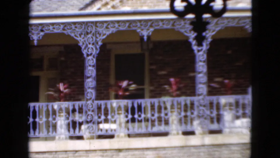 1952 Florida home-movie frame of a brick balcony facade with white decorative railings, shaded openings, and potted plants.