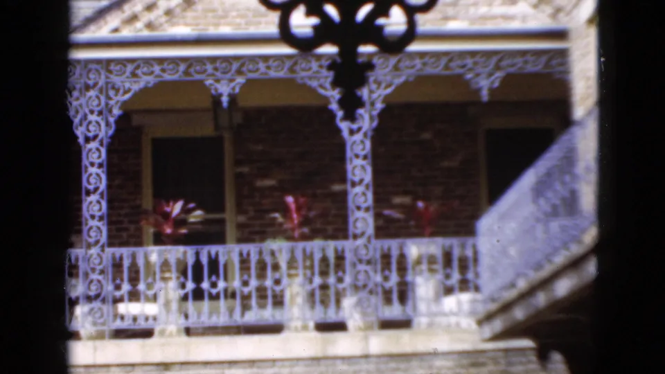 1952 Florida 8mm view with ornate white balcony posts, railing, brick facade, potted red plants, and a partial stair or balcony edge.