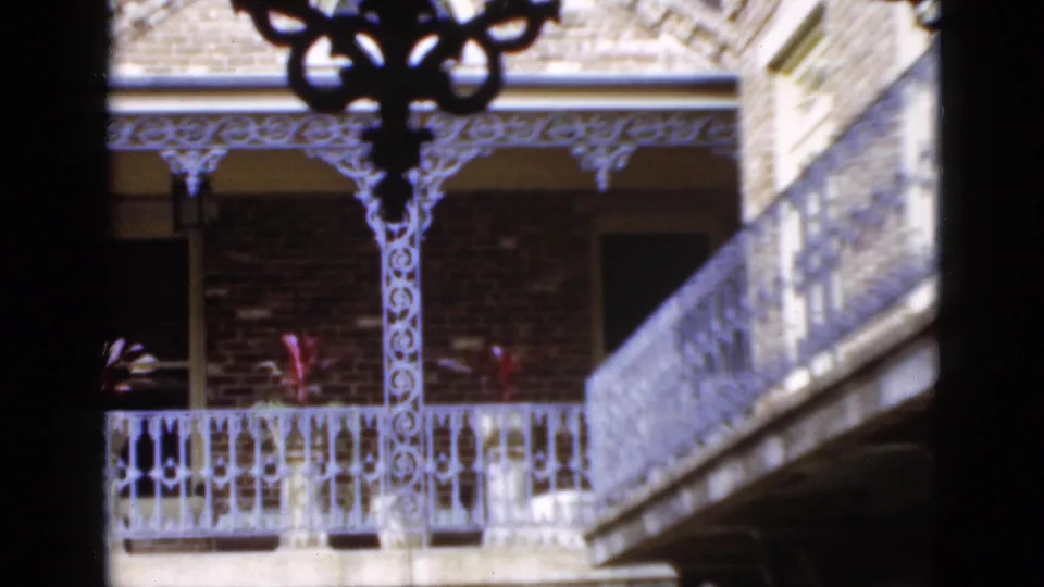 1952 Florida home-movie frame showing decorative balcony ironwork, brick facade, red plants, and a foreground stair or balcony edge.