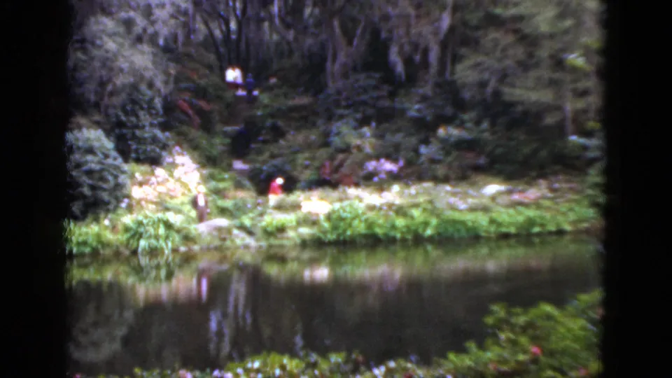 1952 Florida home-movie frame looking across still water toward a lush garden bank and a distant person in red.
