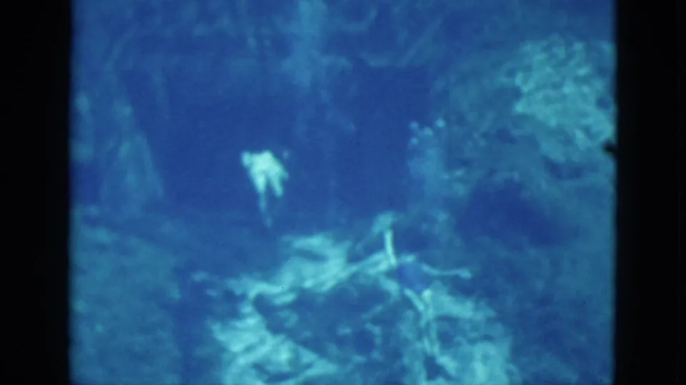 1952 Florida underwater home-movie frame with a distant swimmer in pale swimwear near rocky or coral-like seabed forms.