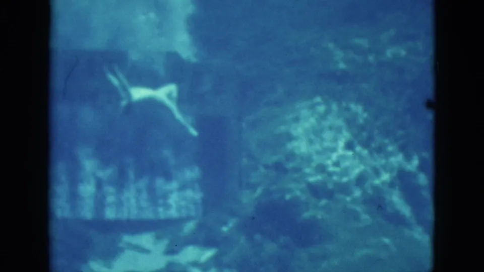 1952 Florida underwater home-movie frame with a swimmer moving past textured rock or reef forms in blue-tinted water.