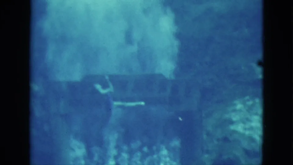 Grainy 8mm frame of a swimmer mid-turn underwater in Florida, with blue-green water and soft cloudlike exposure above.