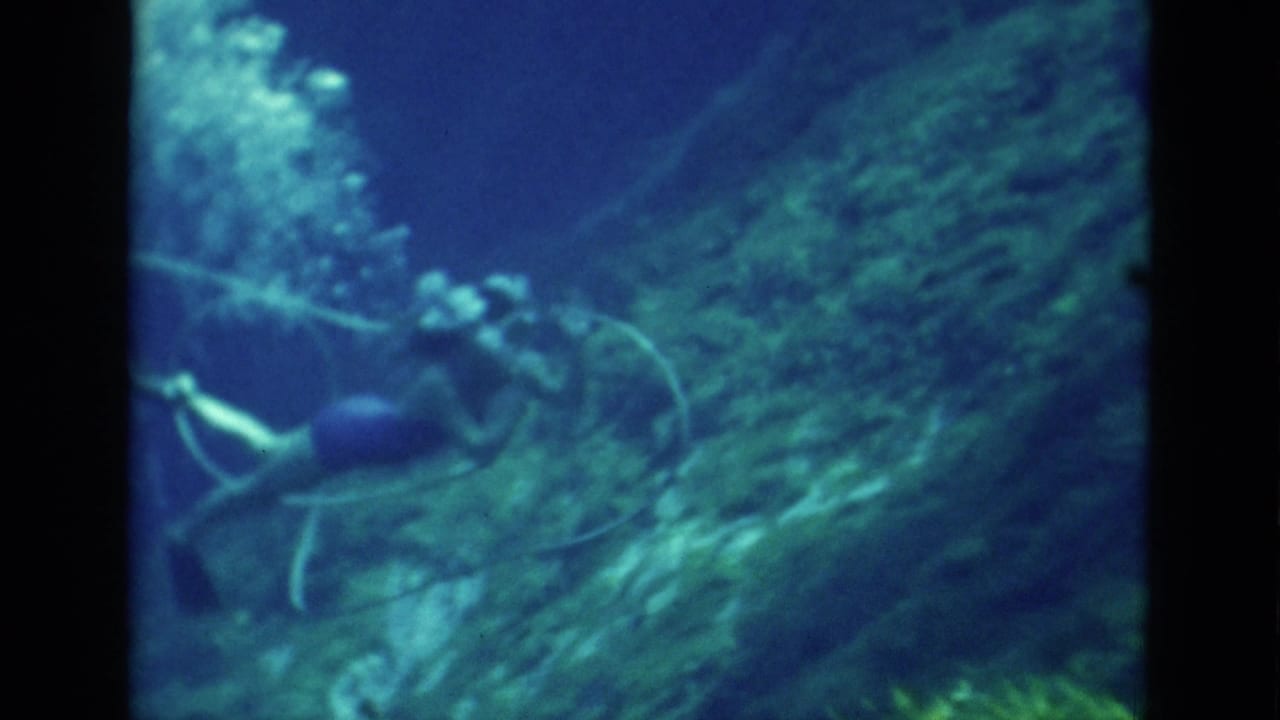 Scuba diver swims along a rocky underwater reef with bubbles rising behind.
