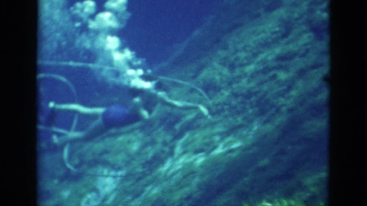 1952 Florida 8mm frame showing a scuba diver from behind beside reef-like rock, with a visible plume of bubbles and trailing hose.
