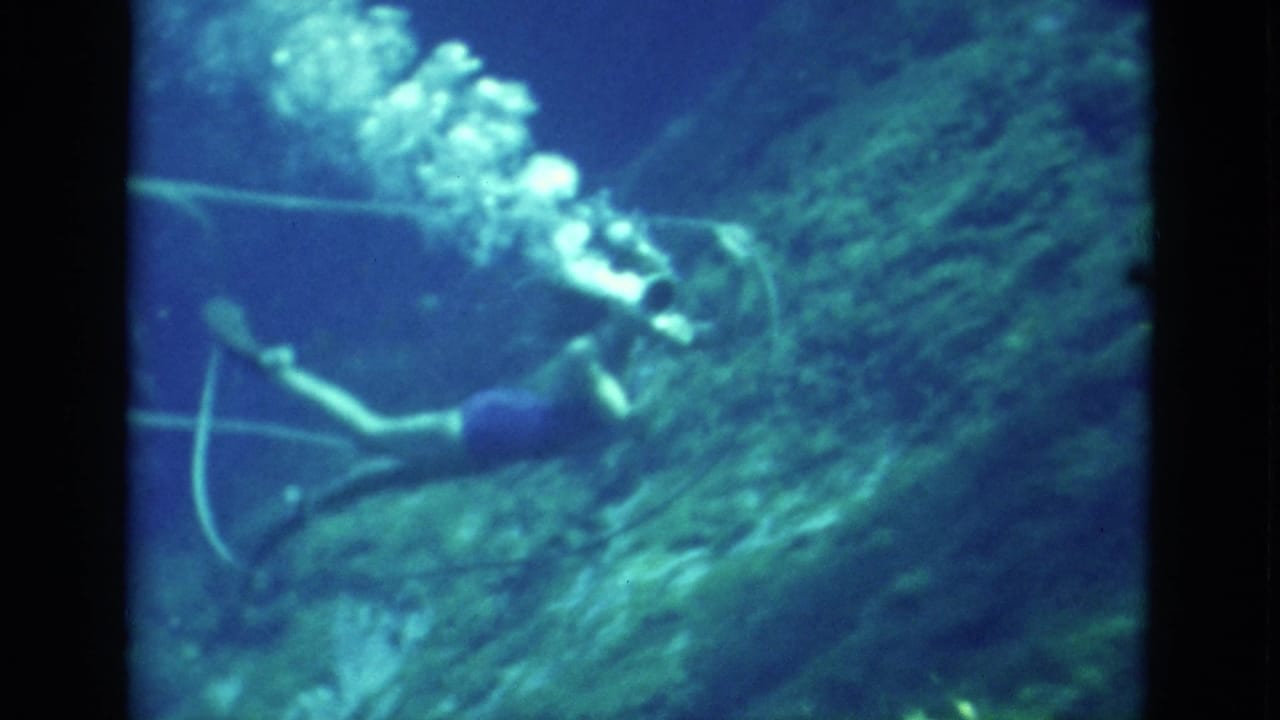 1952 Florida 8mm frame of a diver swimming past reef-like rock and marine growth, with bubbles drifting upward in blue-green water.