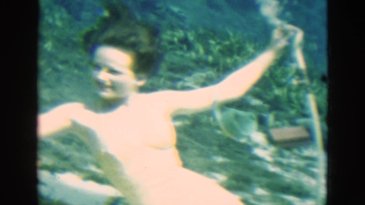 Adult swimmer faces the camera underwater in Florida, one arm raised near a snorkel tube amid blue-green water and plant growth.