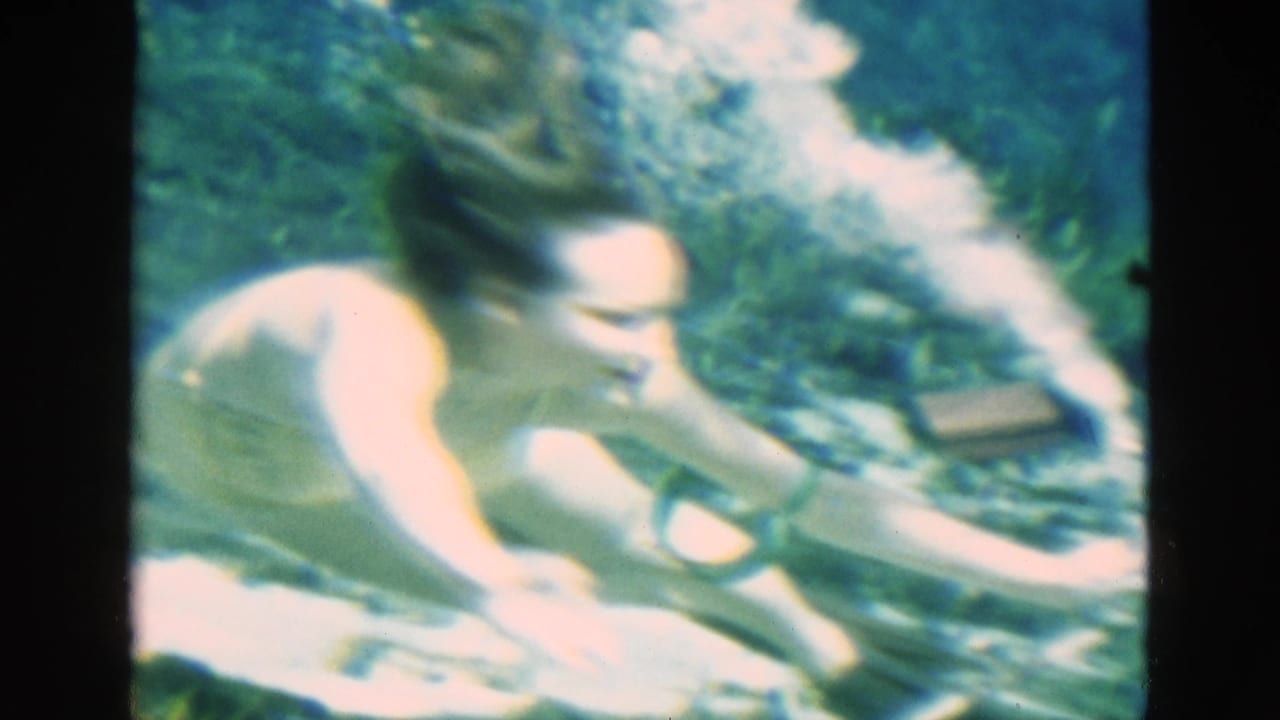 A woman in a vintage swimsuit bends forward underwater near rocks and plants while the breathing tube remains close beside her.