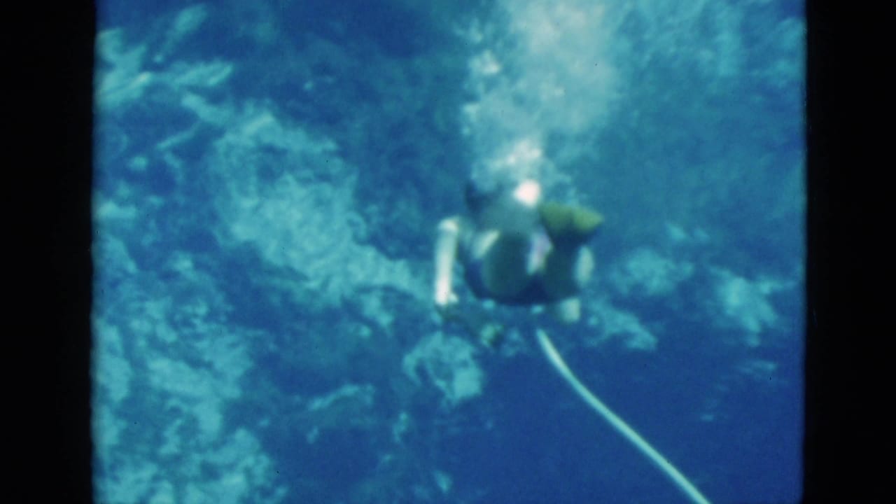 1952 Florida 8mm frame of a woman underwater with an oxygen hose, seen from above against blue water and rocky formations.