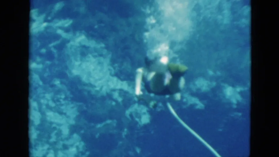 1952 Florida 8mm frame of a woman underwater with an oxygen hose, seen from above against blue water and rocky formations.