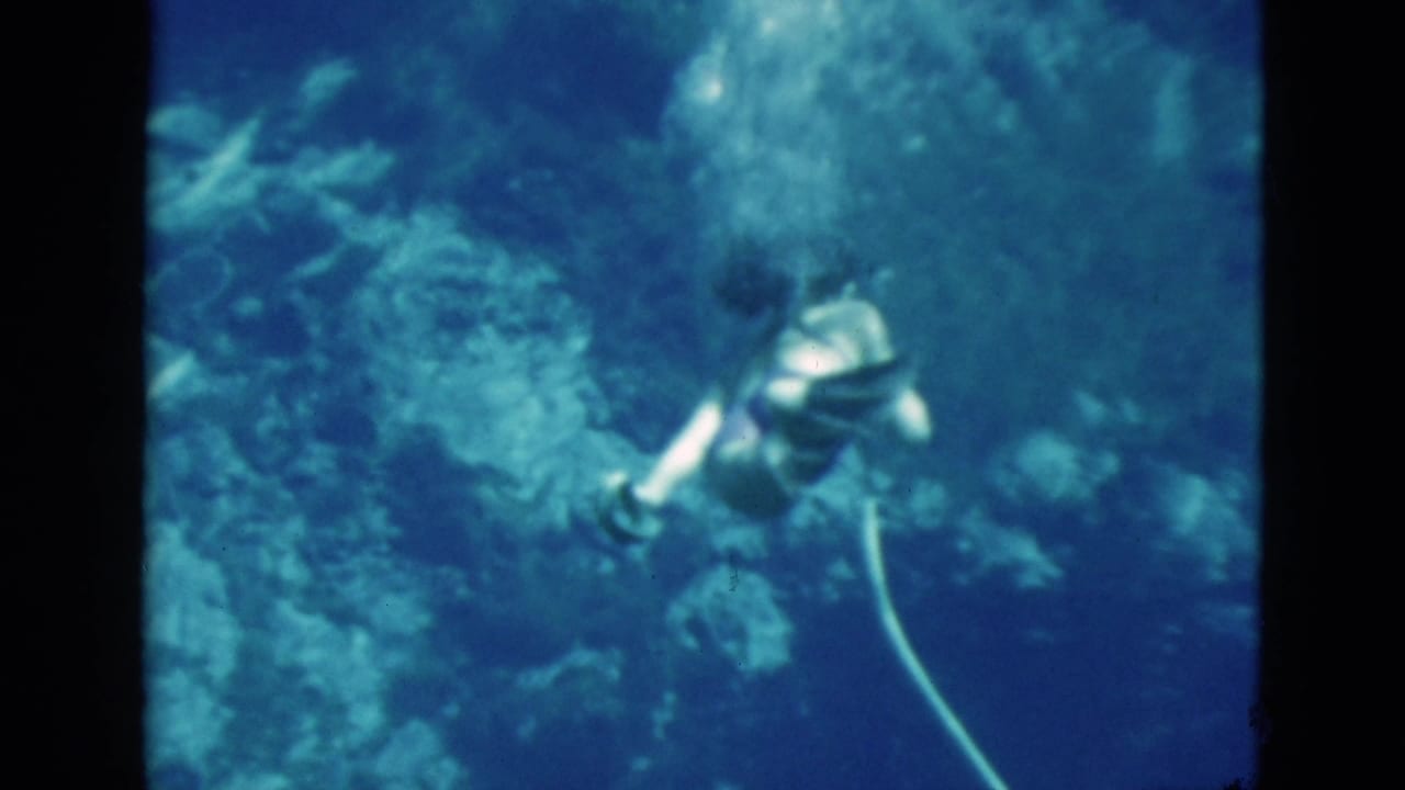 Swimmer underwater with hose near rocky formations in blue water.