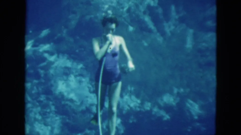 A woman in a purple one-piece swimsuit faces the camera underwater in 1952 Florida footage, with a hose rising beside her.