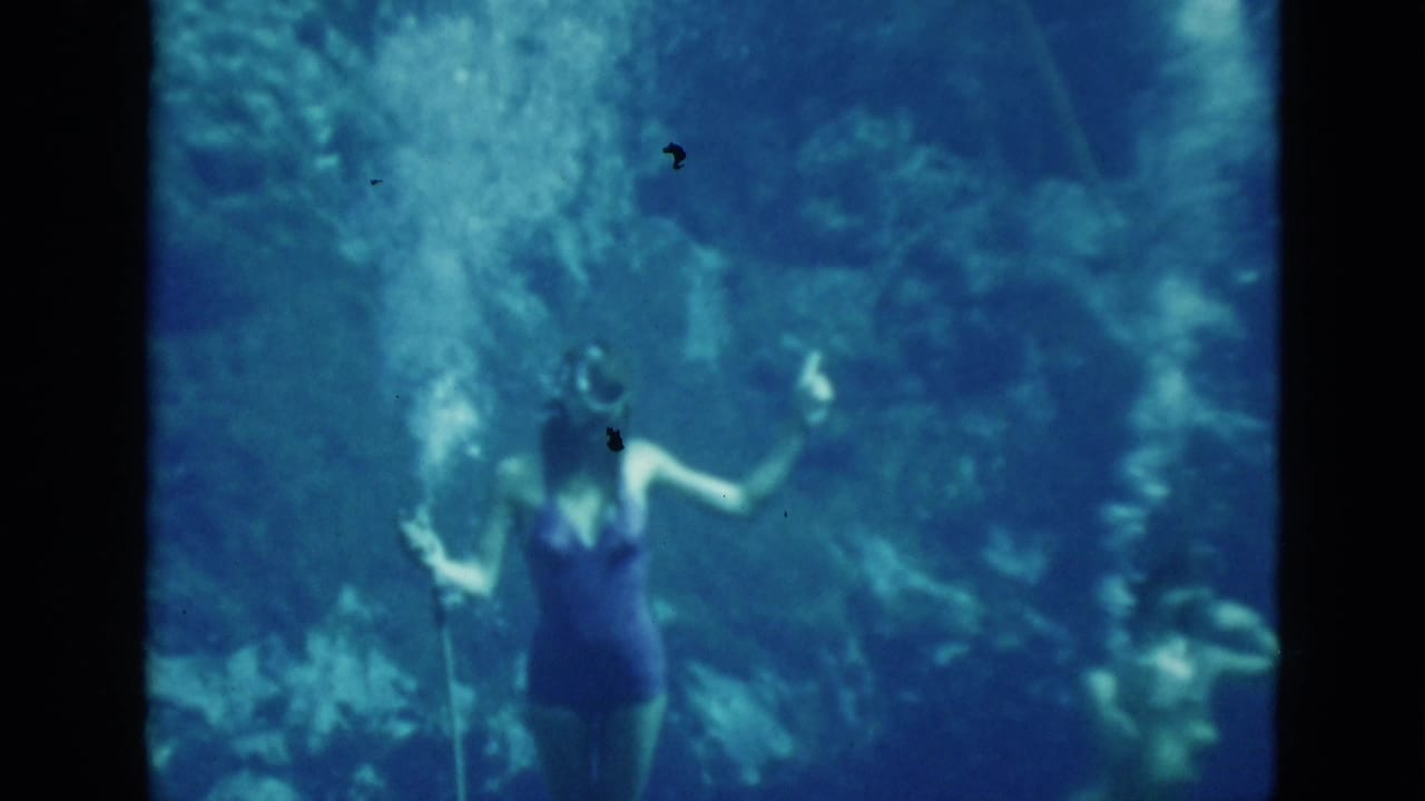 A woman in a dark one-piece swimsuit holds an air hose underwater.