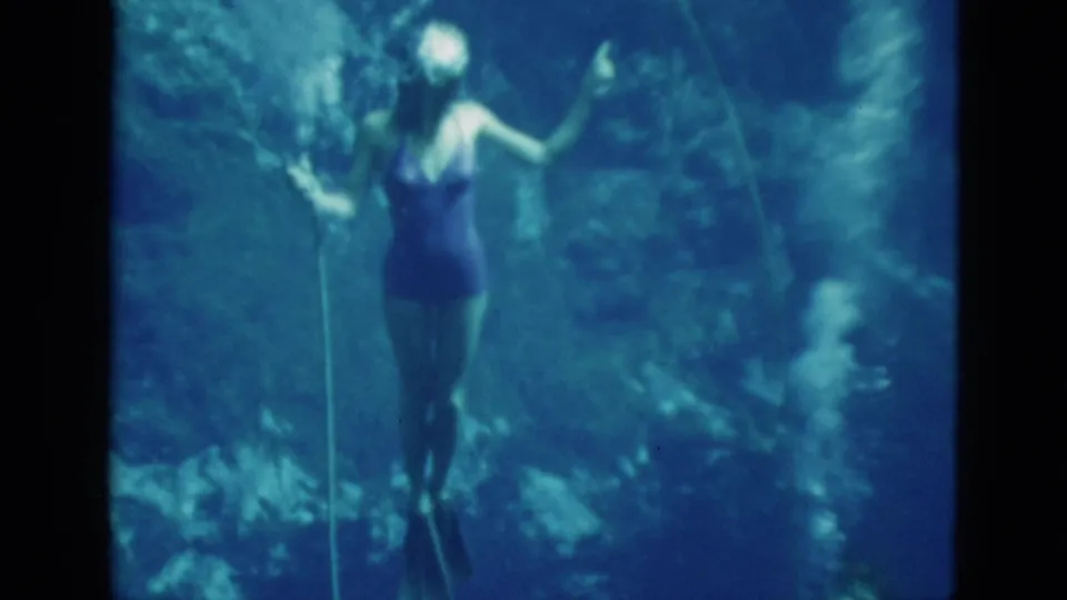 A young woman with fins and a one-piece swimsuit appears underwater beside a vertical air hose in the blue 8mm frame.