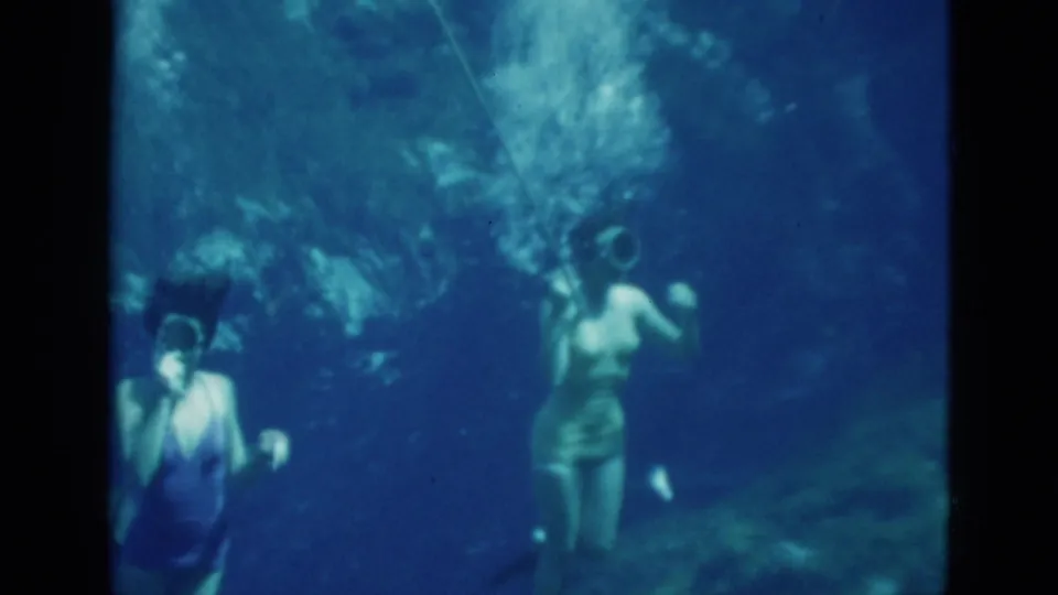 Two young women are visible underwater, one nearer the camera and another swimming away with fins in blue Florida 8mm footage.