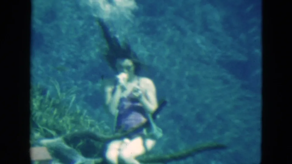 1952 Florida home-movie frame showing an underwater woman in a purple swimsuit holding a breathing tube close to her body as bubbles rise above.