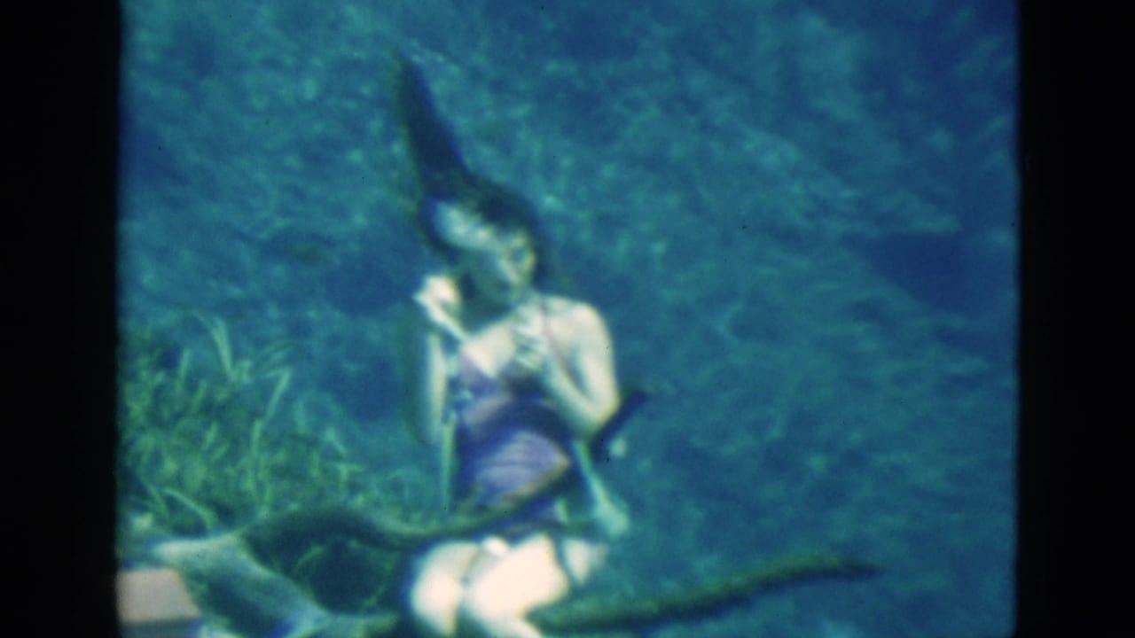 1952 Florida 8mm frame showing a young woman swimming underwater in blue-green water near aquatic plants and dark branches.