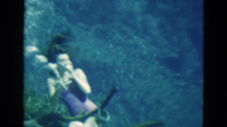 1952 Florida 8mm frame showing a woman underwater with hands near her face, surrounded by blue-green water and faint plant growth.