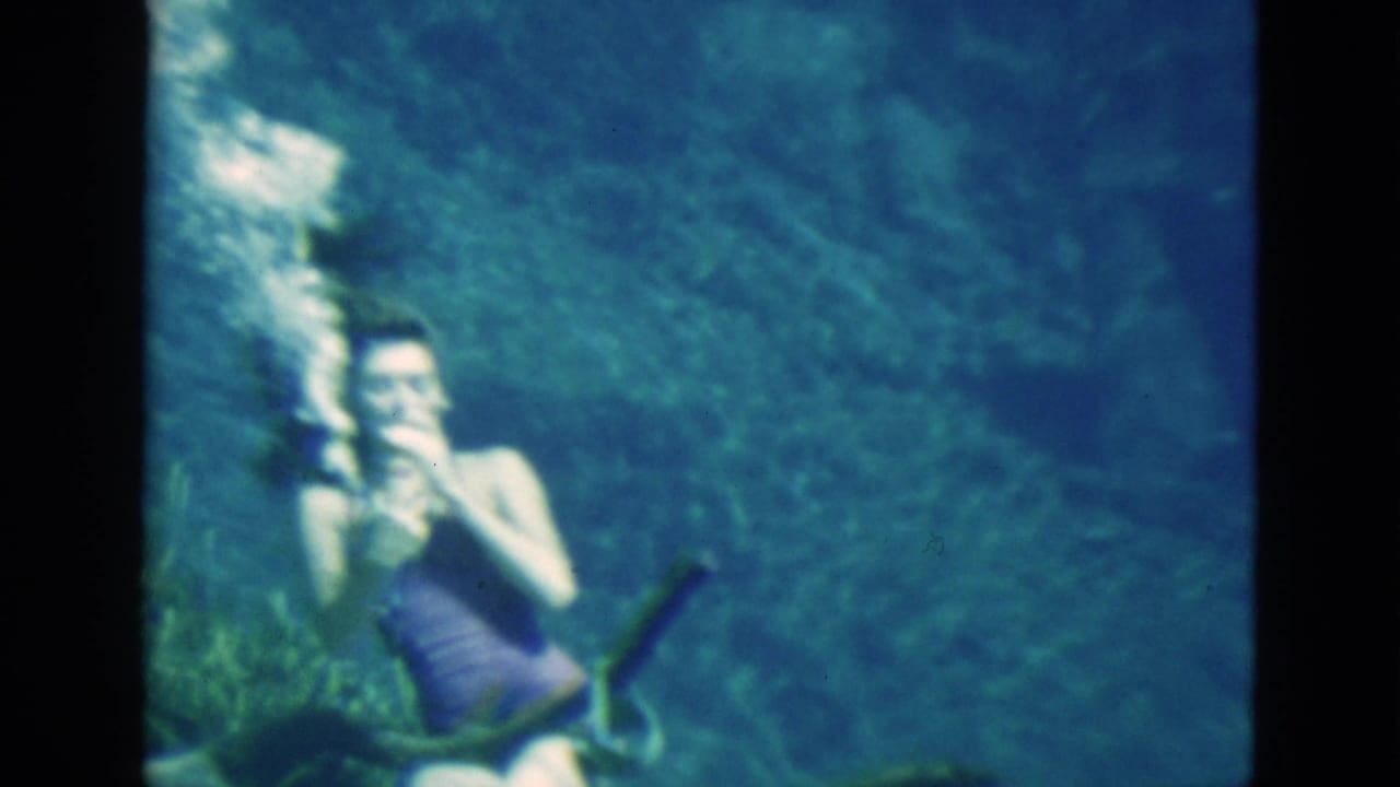 1952 Florida 8mm frame of a woman underwater in a purple swimsuit, with bubbles around her as she handles a mask and bottle.