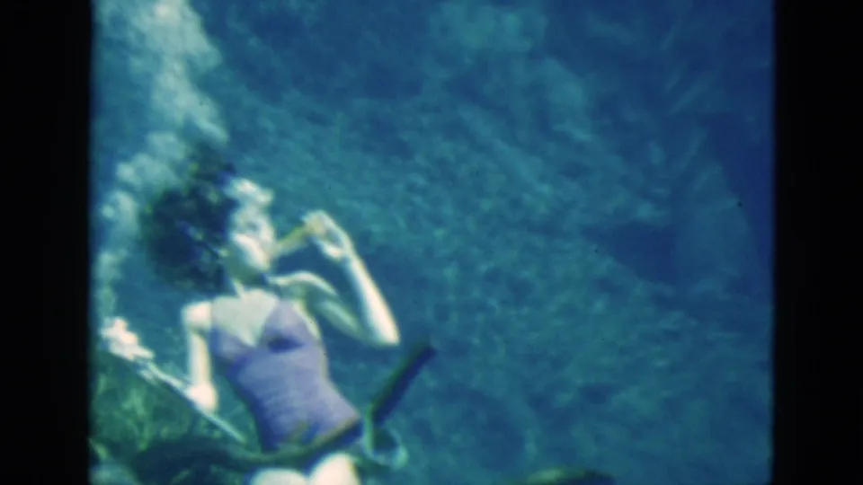 1952 Florida home-movie frame showing a woman underwater in a purple swimsuit raising a bottle near her mouth.