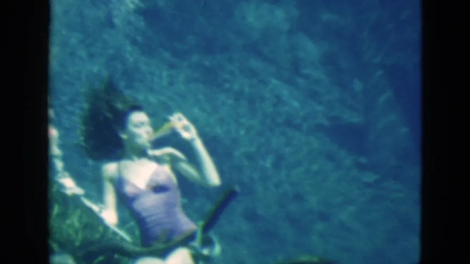 1952 Florida 8mm frame of a woman in a swimsuit underwater, holding a bottle near her mouth with gear across the foreground.
