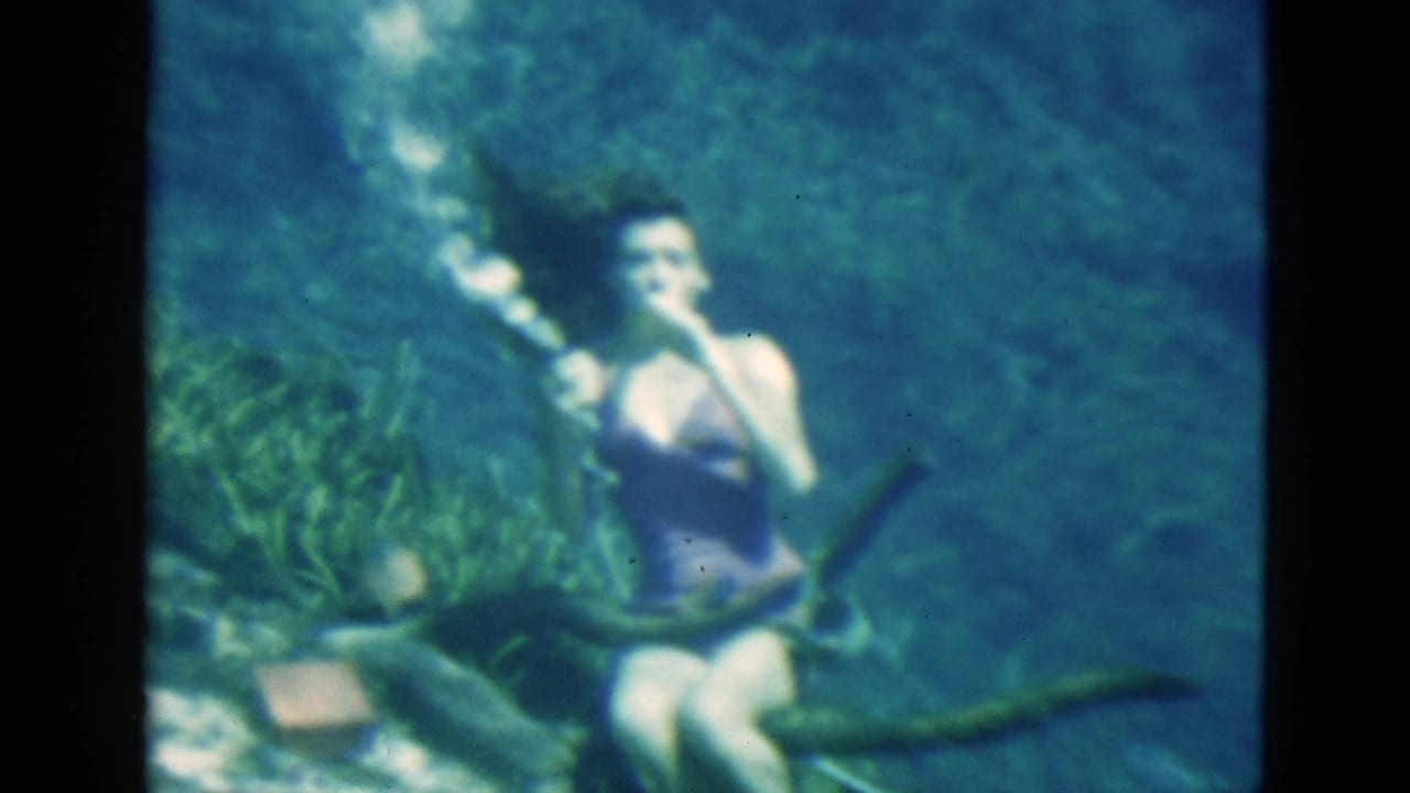 1952 Florida 8mm frame of a woman in a purple swimsuit underwater near aquatic plants, holding a glass as bubbles rise.