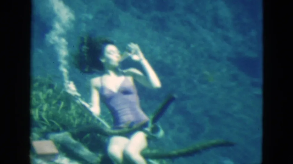 1952 Florida 8mm frame of a woman drinking underwater in a clear spring or lake, seated beside aquatic plants and a textured bottom.
