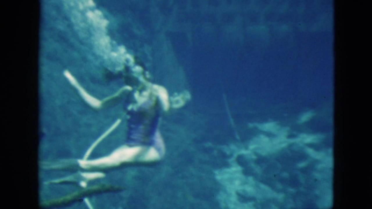 1952 Florida 8mm frame of a woman swimming underwater in a purple swimsuit, with blue water, rocks, bubbles, and a pale hose or snorkel line.