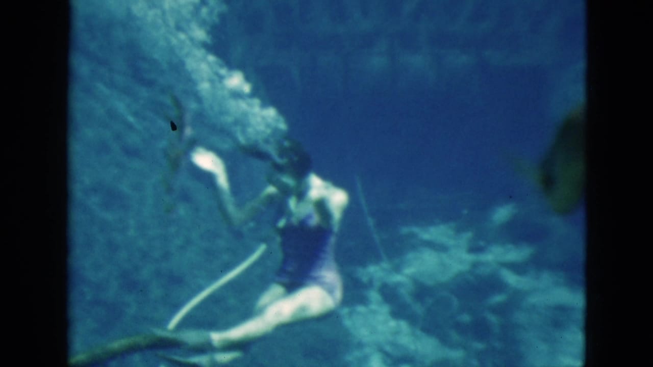 1952 Florida home-movie frame showing a woman underwater in a purple swimsuit, with bubbles, rocky formations, and soft 8mm color shift.