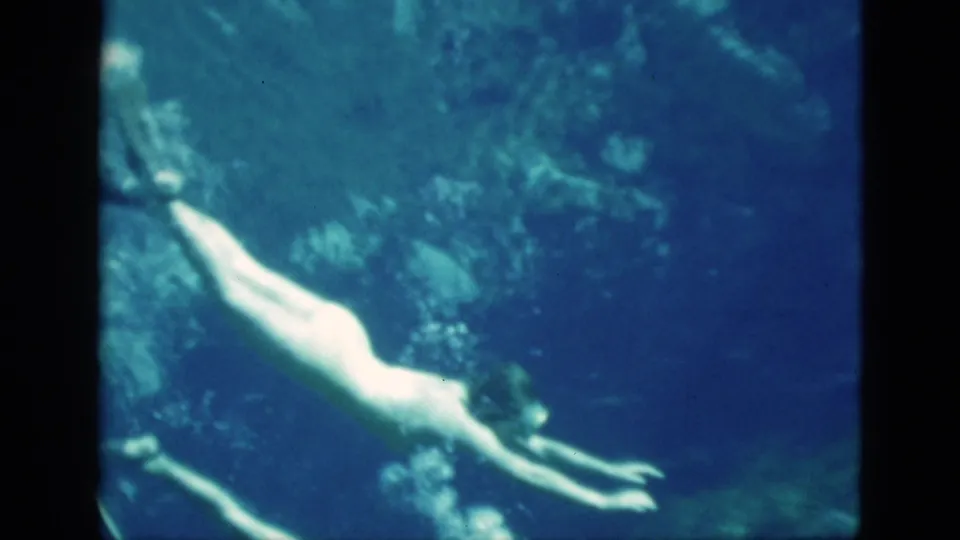 1952 Florida 8mm frame of a woman swimming underwater in a swimsuit and fins, with blue-green water and aquatic background visible.