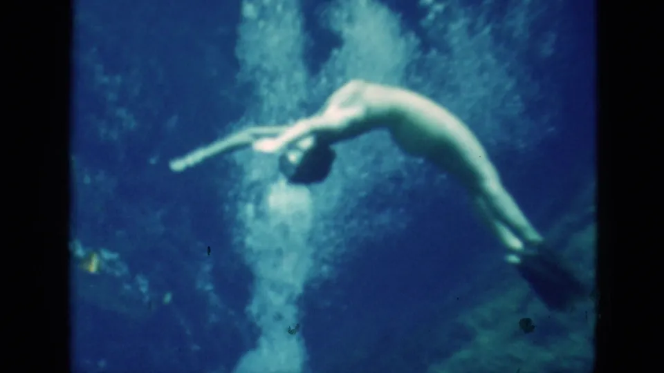 1952 Florida 8mm frame of a woman curving downward underwater in a swimsuit and fins, surrounded by bright bubbles.