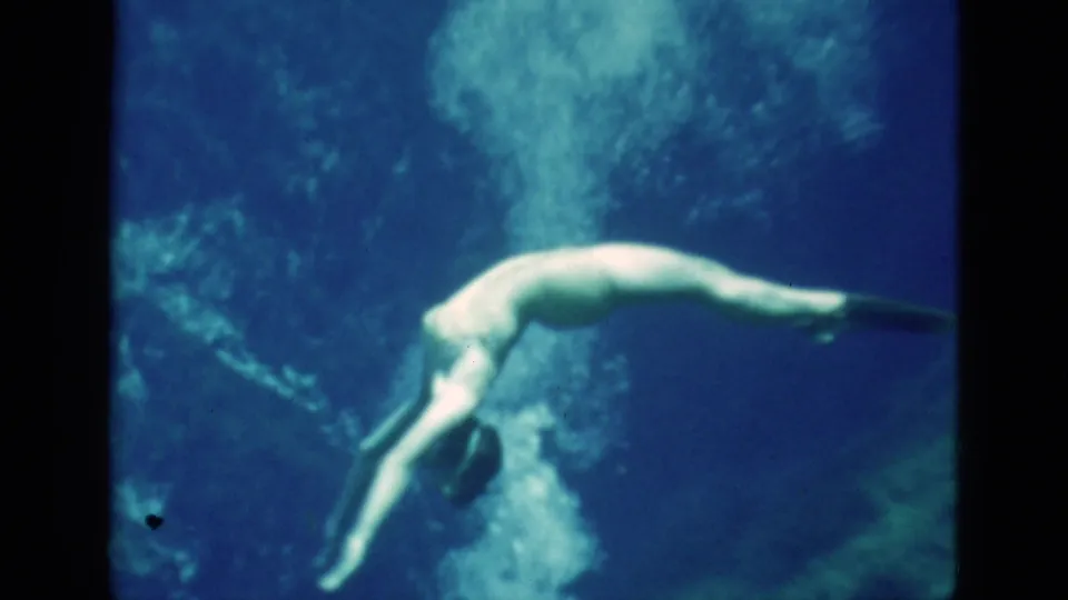 1952 Florida 8mm frame of a woman underwater, angled downward with swim fins as bubbles rise behind her in blue water.