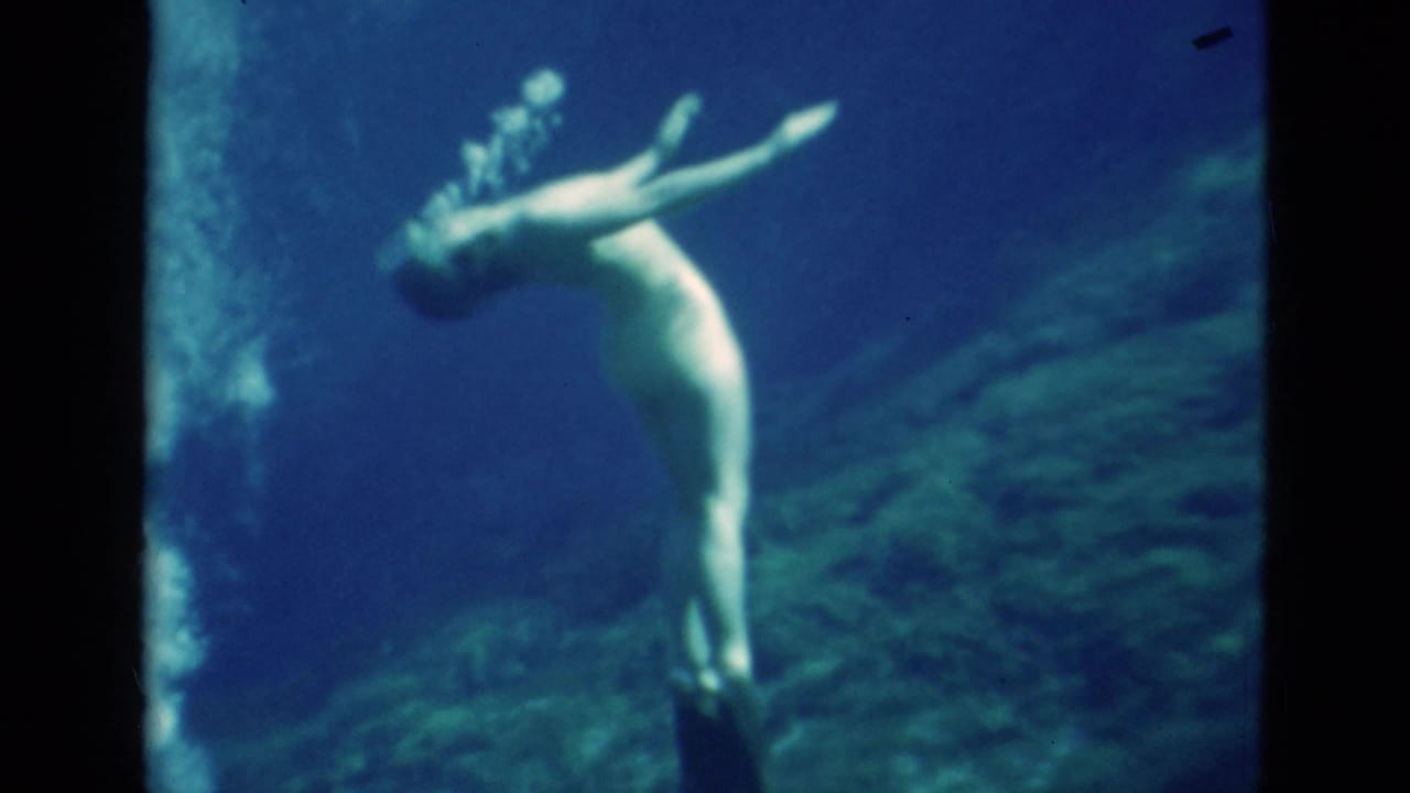1952 Florida 8mm frame showing a woman underwater in an upright pose with arms lifted and bubbles rising in the surrounding blue water.